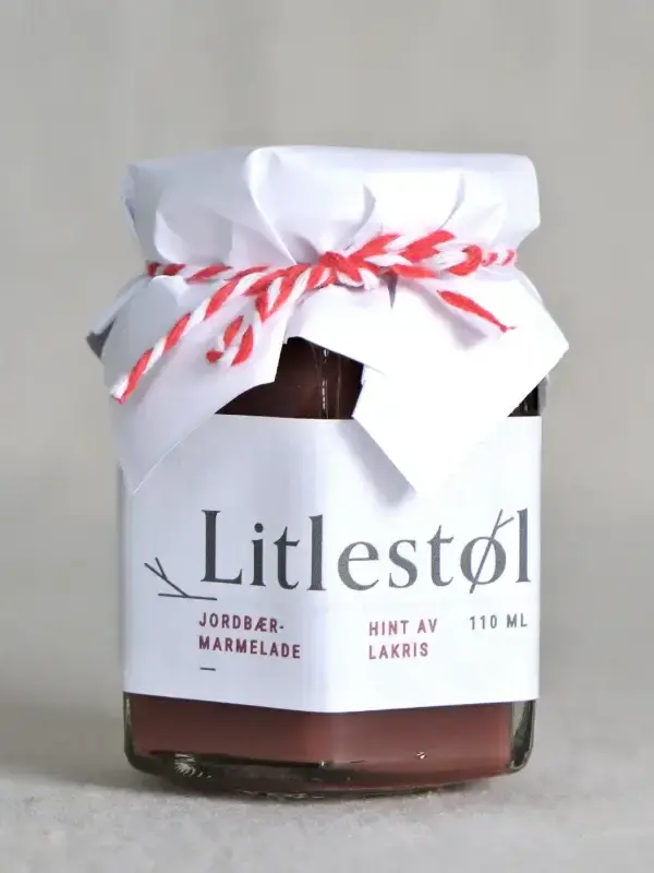 Glass med Litlestøl jordbærmarmelade med lakris og lime 110ml, plassert på et trebord med ost, nøtter og ferskt brød.