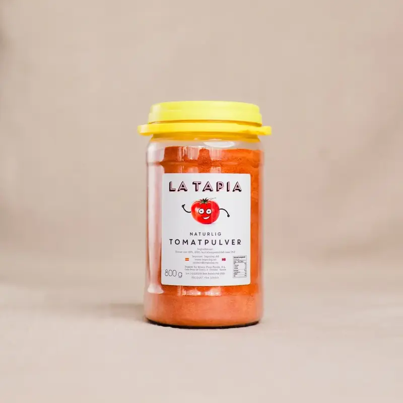 Beholder med La Tapia tomatpulver, plassert ved siden av frisk tomat på kjøkkenbenken.