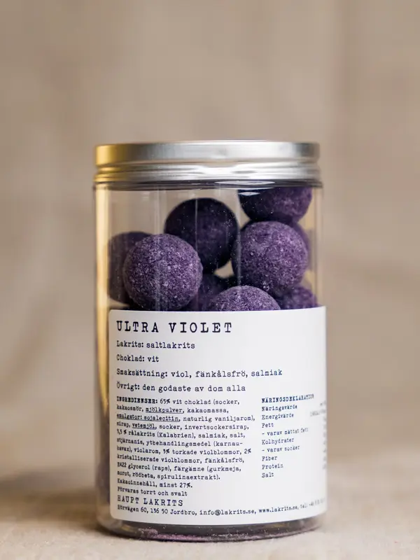 Et glass med Ultraviolet fra Haupt – lakriskuler med en kjerne av langkokt saltlakris, trukket i hvit sjokolade smaksatt med viol, salmiakk og fänkålsfrø, presentert i en elegant glasskrukke med etikett.