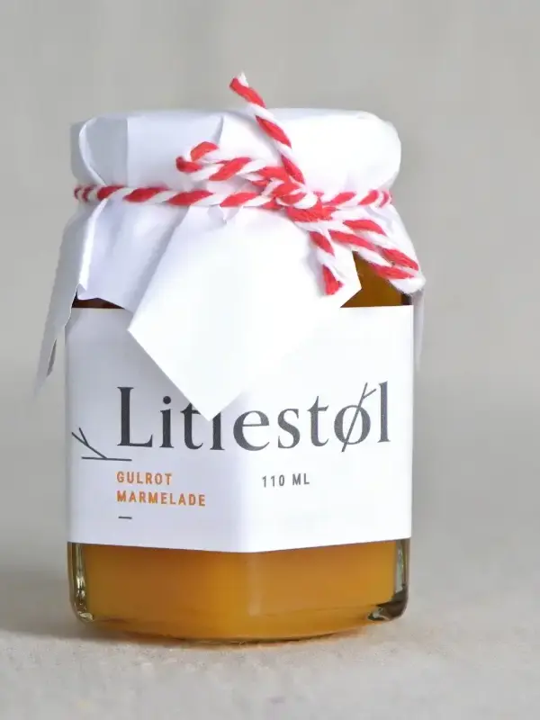 Glass med Litlestøl gulrotmarmelade 110ml, plassert på et trebord med ost, nøtter og ferskt brød.