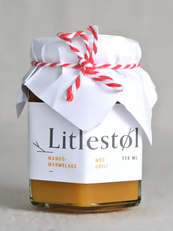 Glass med Litlestøl mango- og chilimarmelade 110ml, plassert på et trebord med ost, nøtter og ferskt brød.