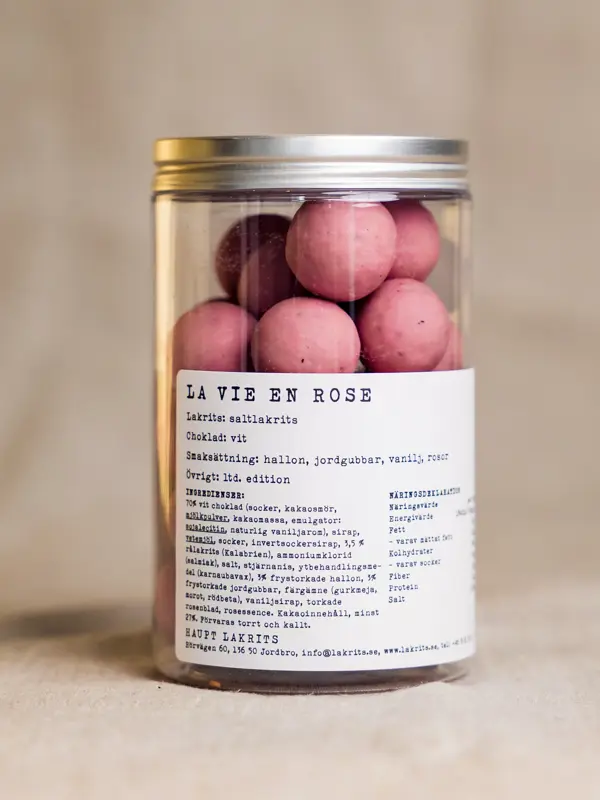 Et glass med La Vie en Rose fra Haupt – rosa lakriskuler med salt lakris kjerne, trukket i hvit sjokolade smaksatt med bringebær, jordbær, vanilje og roseblader, presentert i en elegant glasskrukke med etikett.