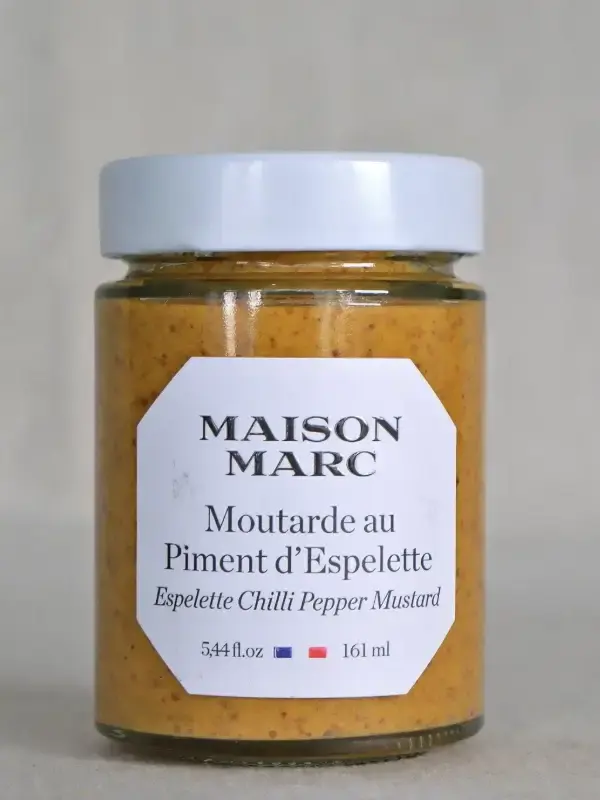 Glass med Dijon sennep fra Maison Marc, smaksatt med Piment d'Espelette, plassert på et bord med friske urter og baguette.